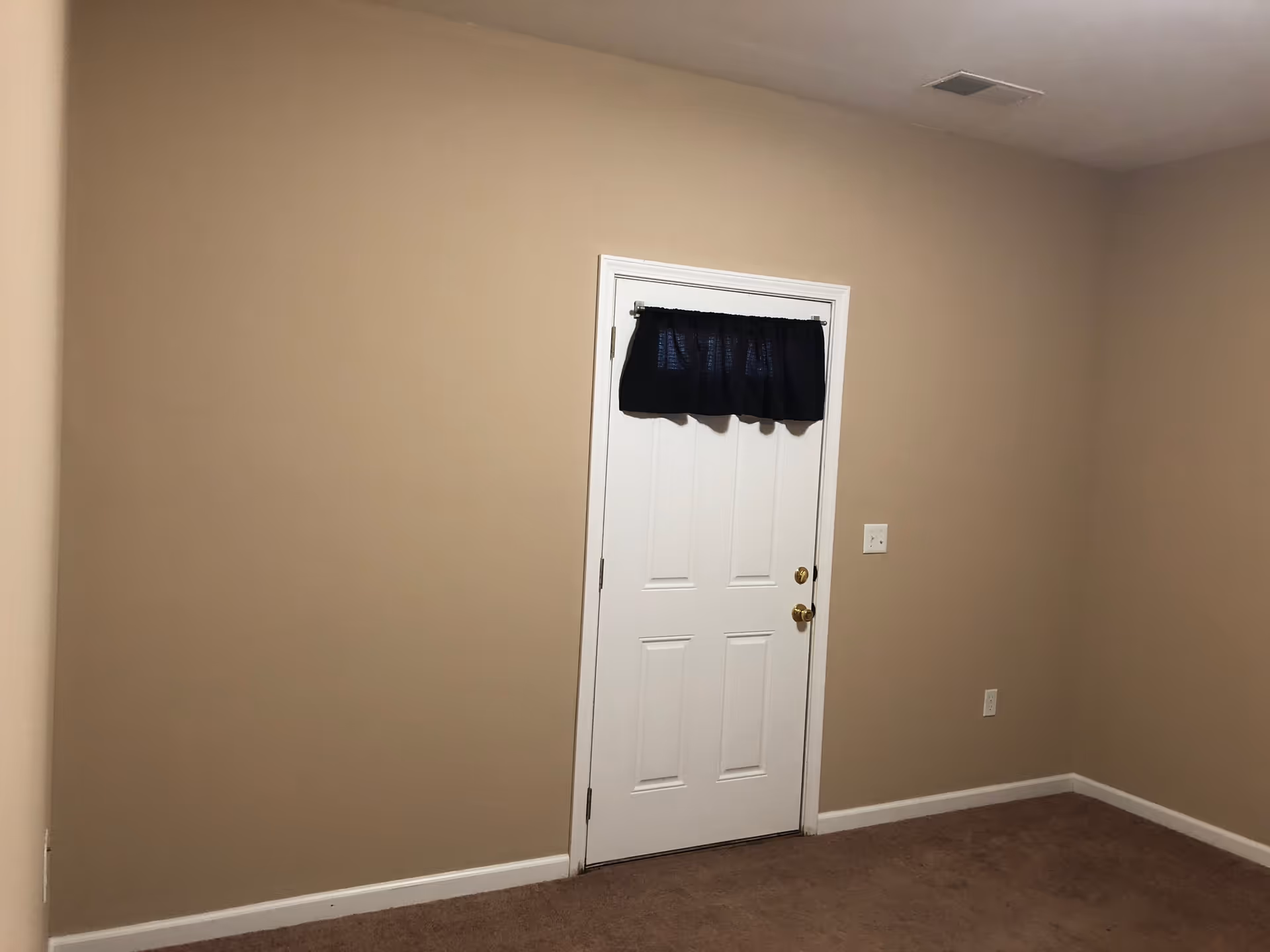 Interior room with beige walls, brown carpet, and a white door with a small dark curtain.