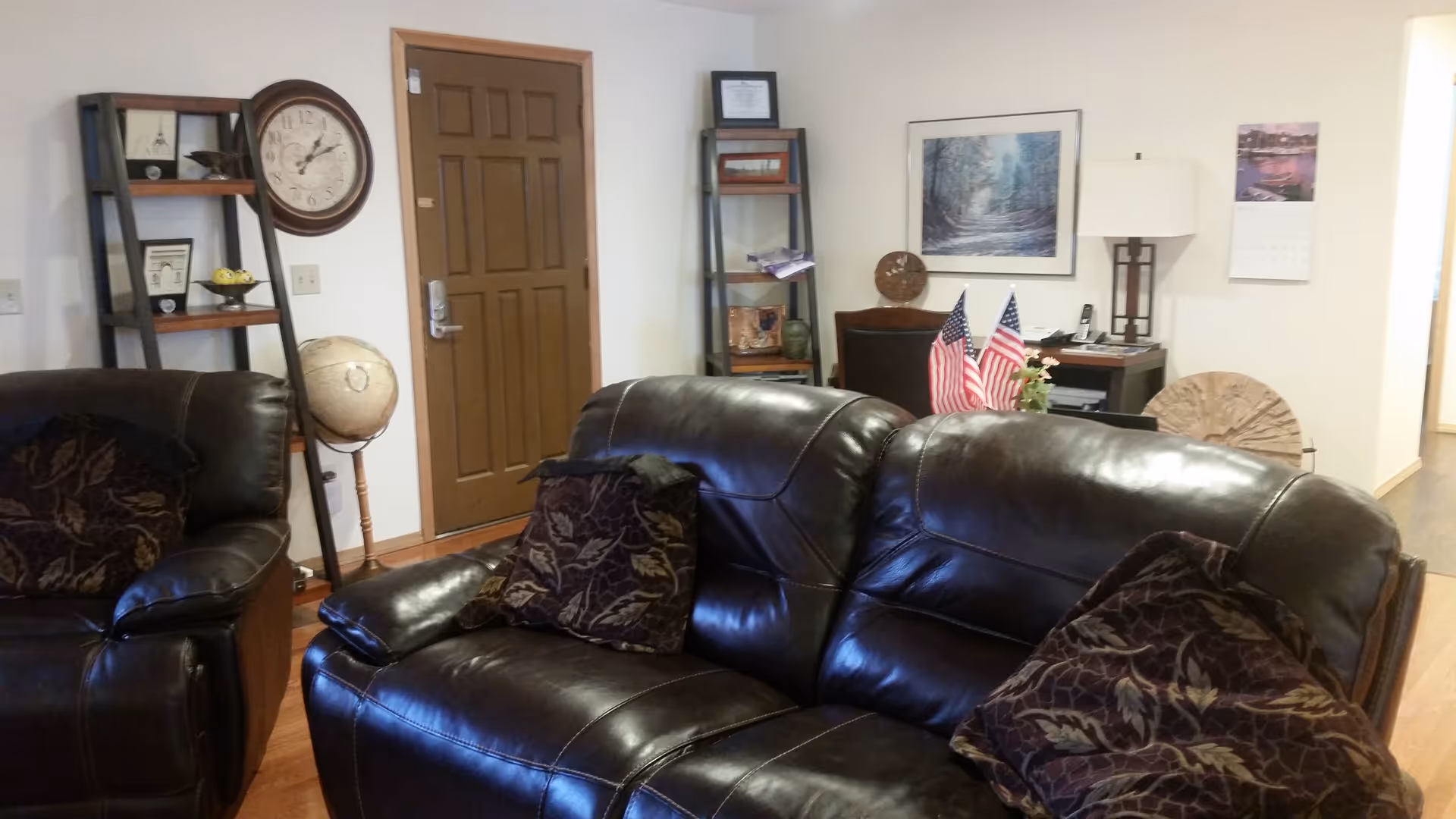 Interior view of a senior living facility common area with dark brown leather sofas and patterned cushions. In the background, there is a wooden door, a wall clock, two shelving units with decorative items, a globe, a desk with a lamp, two small American flags, a calendar, and framed artwork on the wall.