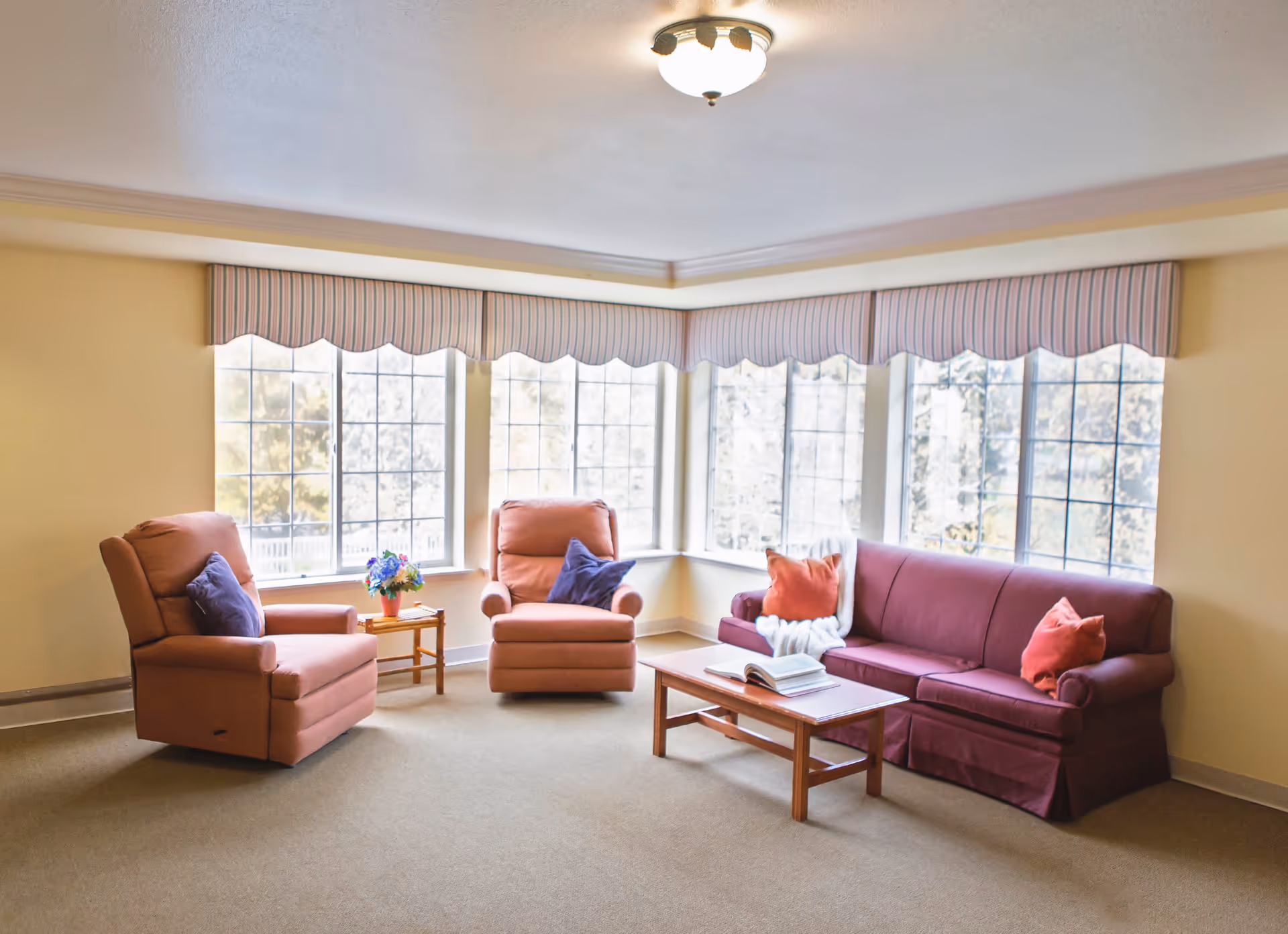 A cozy living room with two peach-colored armchairs and a maroon sofa adorned with orange and purple pillows. A wooden coffee table with an open book sits in front of the sofa. Large windows with striped valances let in natural light, illuminating the beige carpet and cream-colored walls.