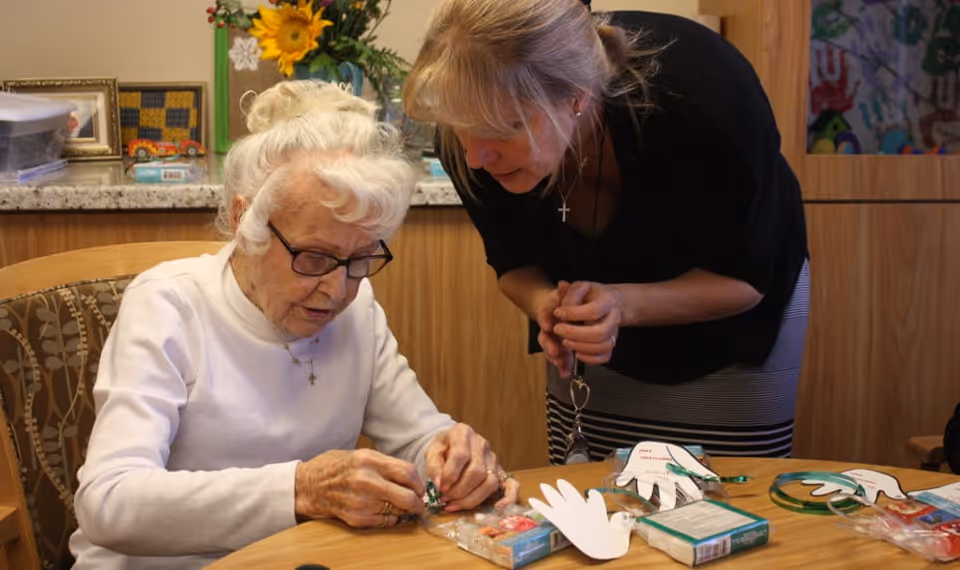 An elderly woman with white hair and glasses sitting at a table, engaged in a craft activity with a younger woman standing beside her, assisting. The table has craft materials including paper cutouts shaped like hands and other supplies. The background shows a wooden cabinet and decorative items.