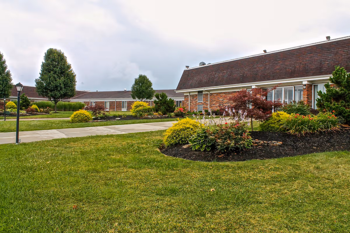Front exterior of a single-story brick senior living building with manicured lawns, flowerbeds, and a paved walkway under a cloudy sky.