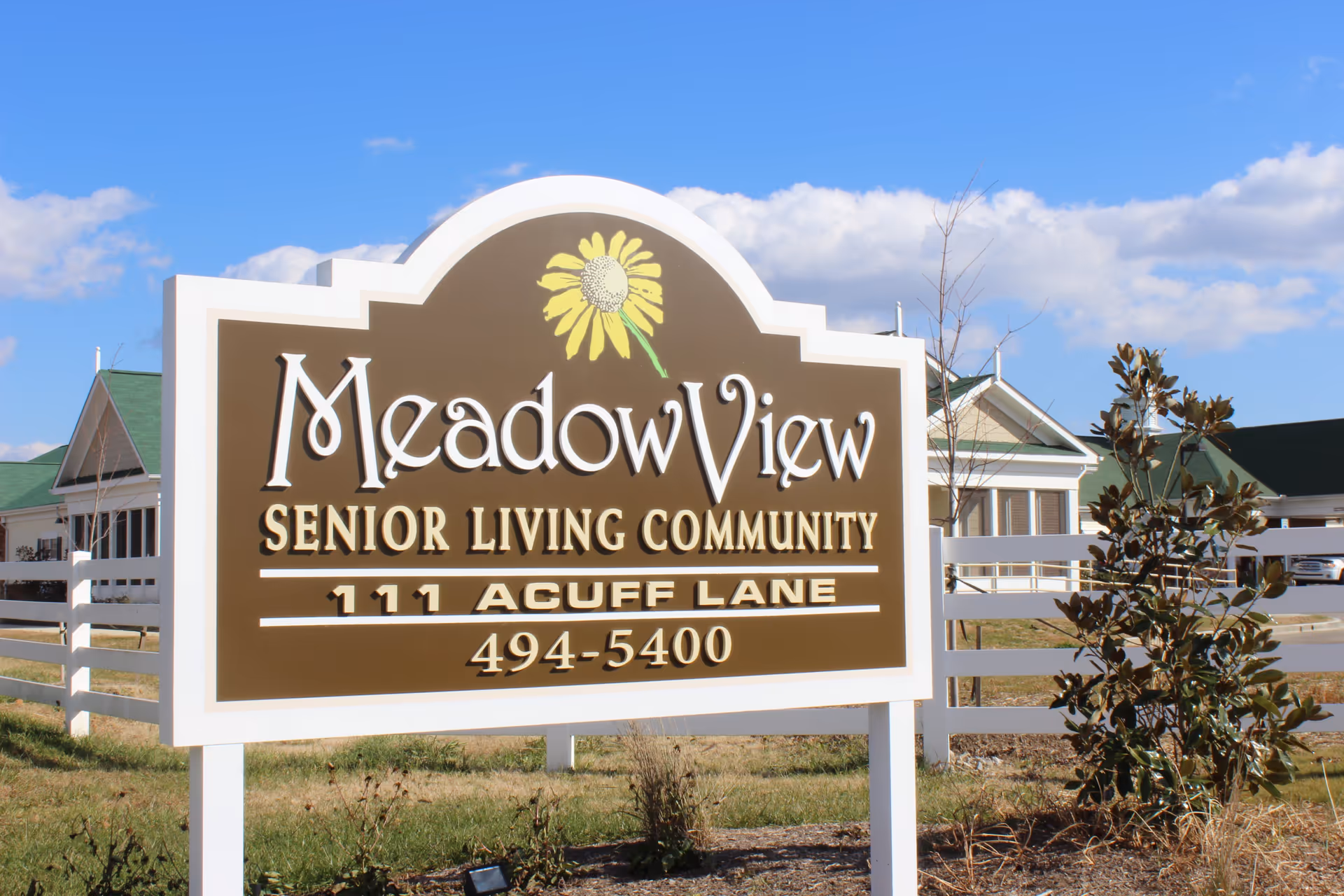 A large outdoor sign for Meadow View Senior Living Community with a yellow flower graphic above the name. The sign includes the address 111 Acuff Lane and a phone number 494-5400. The background shows part of the senior living facility buildings, a white fence, some bushes, and a blue sky with clouds.