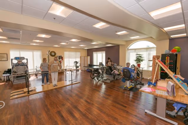 A spacious rehabilitation room with wooden flooring and large windows. The room contains various physical therapy equipment including parallel bars, exercise bikes, and other therapy tools. Two people are using the parallel bars for walking exercises, while others are seated on exercise machines. The room is well-lit with ceiling lights and natural light from the windows.
