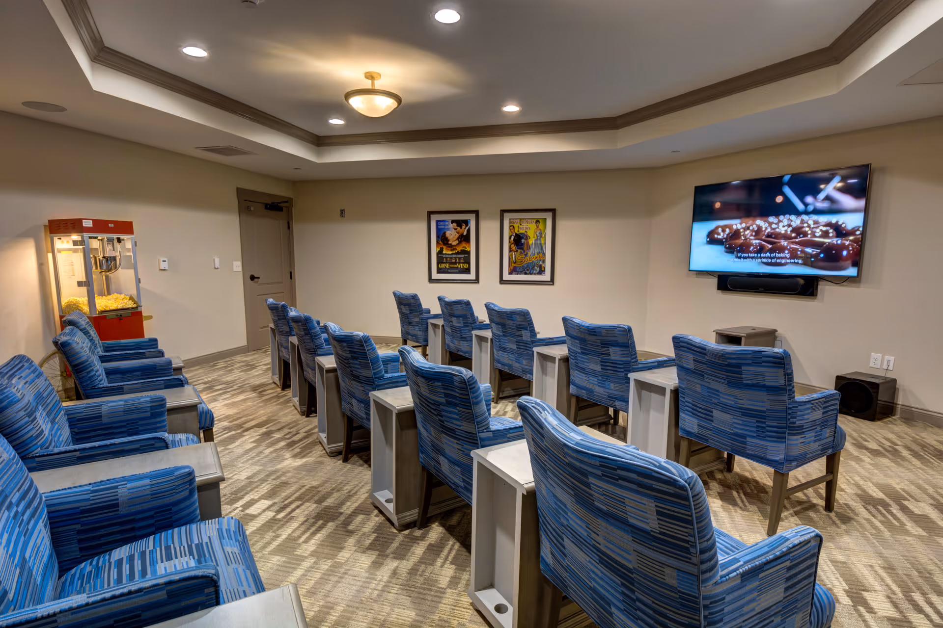 A small movie theater room with multiple rows of blue patterned armchairs facing a wall-mounted flat screen TV. There is a popcorn machine in the corner near the door, and two framed movie posters on the wall behind the chairs. The room has recessed lighting and a carpeted floor.