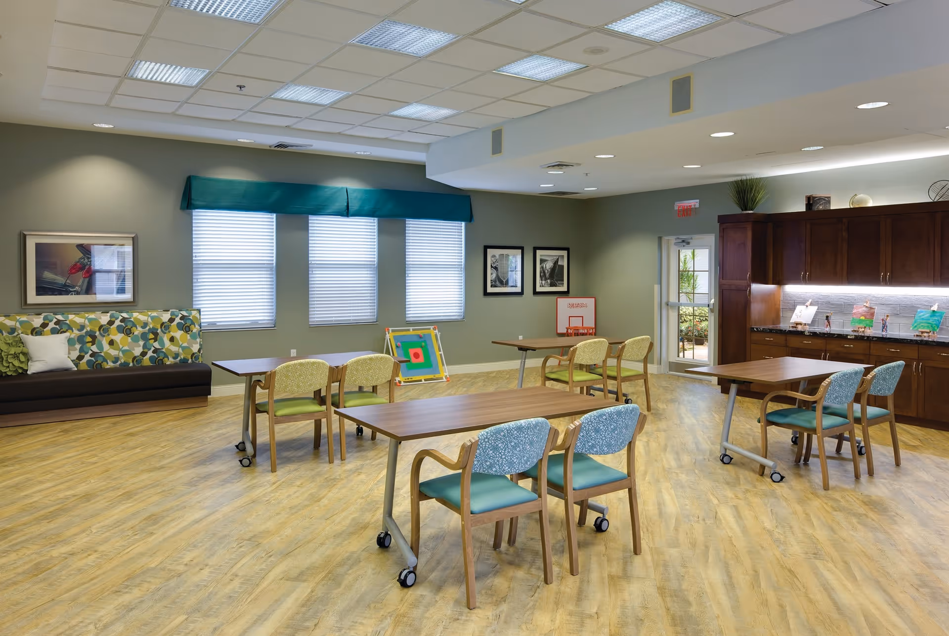 Bright multipurpose activity room with tables and chairs, a couch by windows, and cabinets along the back wall.