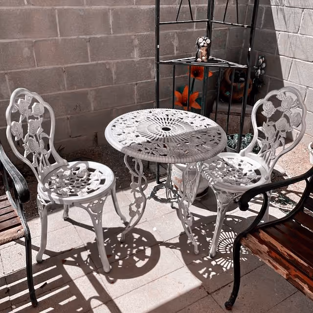Outdoor patio area with a white ornate metal table and two matching chairs, alongside a wooden bench with black metal armrests, set against a concrete block wall with a black metal corner shelf holding decorative items.