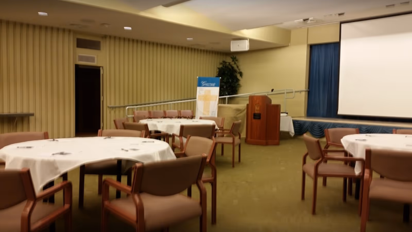 Interior view of a meeting or dining room with round tables covered with white tablecloths and surrounded by brown chairs. There is a wooden podium near a stage with a large white projection screen and blue curtains. A banner with a cross design is visible near the back wall.