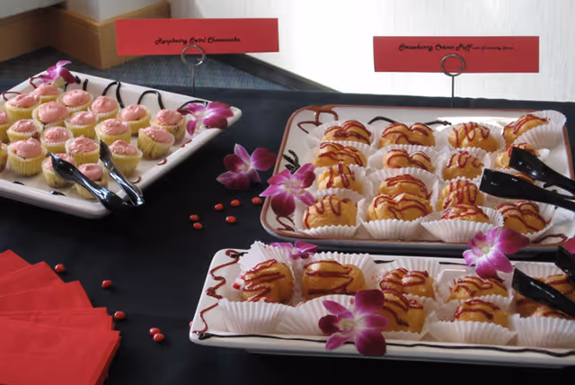 Three rectangular white trays with small desserts arranged neatly on a black tablecloth. The desserts include raspberry swirl cheesecakes and strawberry cream puffs, each placed in white paper cups. Purple orchid flowers are used as decoration on the trays and table. Red napkins and serving tongs are also visible on the table.