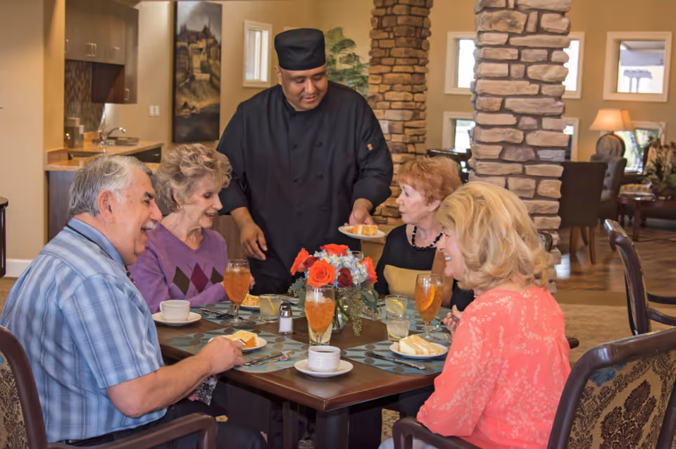 A chef serves plated dessert to four older adults seated around a dining table in a cozy senior living dining room.