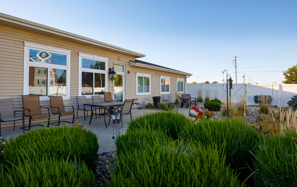 Outdoor patio area at Homestead Assisted Living of Knoxville featuring a row of chairs and a table on a concrete surface, surrounded by green bushes and plants, with a beige building and windows in the background under a clear blue sky.