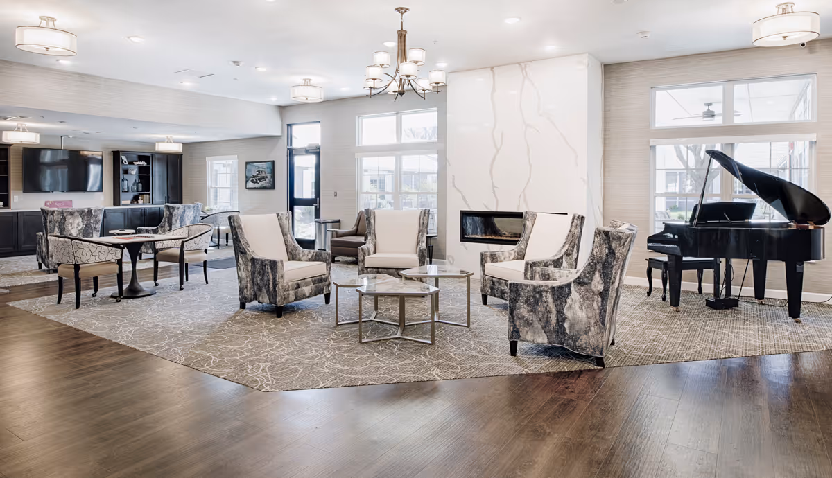 A spacious and well-lit living room area in an assisted living facility featuring a modern fireplace, several upholstered armchairs arranged around a coffee table, a grand piano near large windows, and a television mounted on the wall with built-in shelving and cabinets.