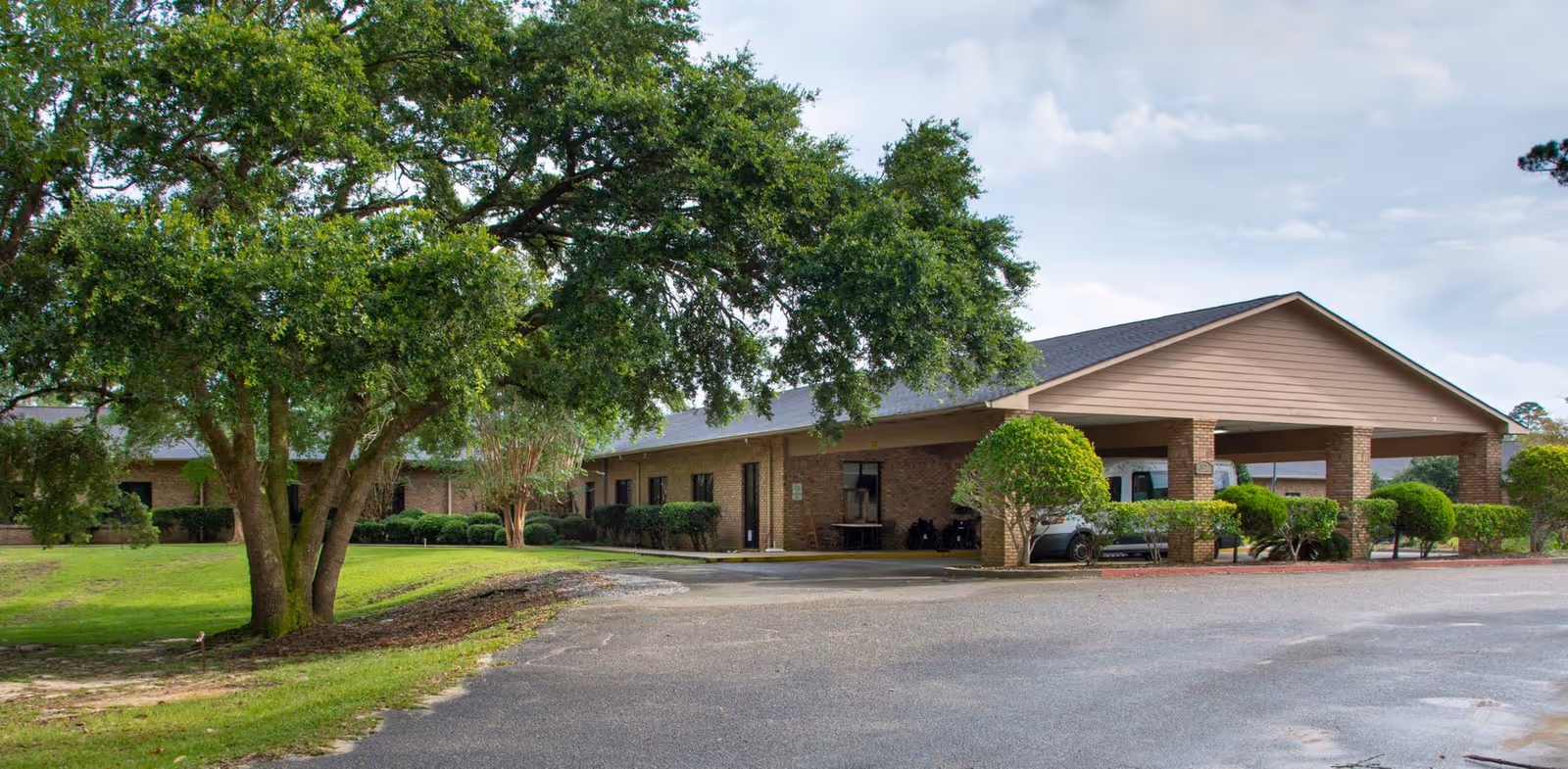 Exterior view of Memorial Woodland Village Rehabilitation and Nursing Center showing a single-story brick building with a covered entrance, surrounded by green trees and shrubs under a partly cloudy sky.