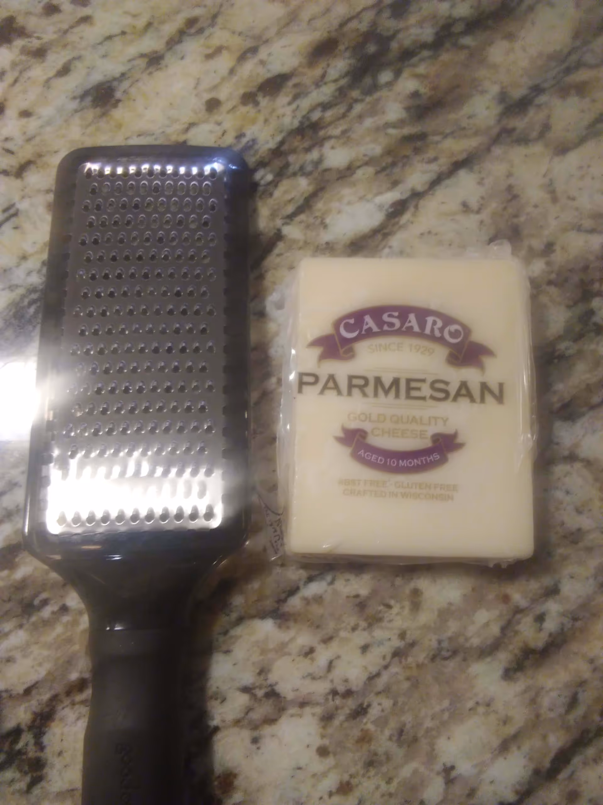 A metal cheese grater with a black handle placed next to a block of Casaro Parmesan cheese on a granite countertop.