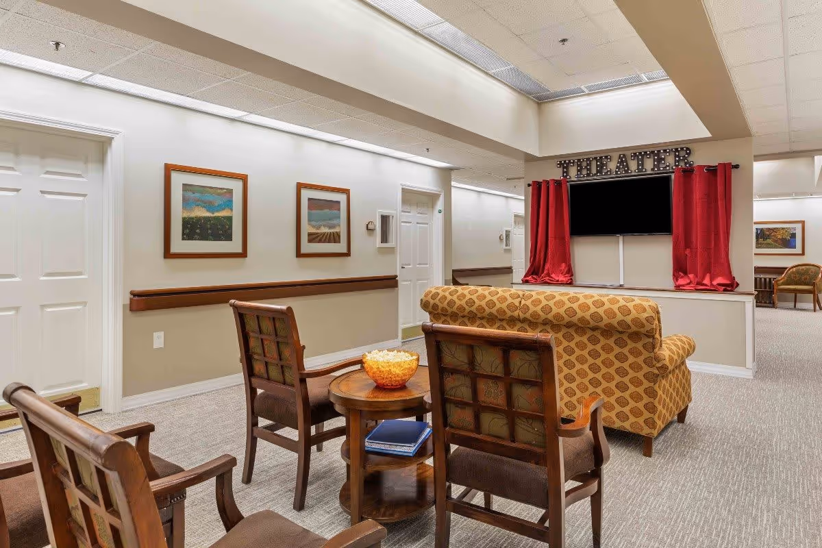 A cozy seating area in a senior living facility with a patterned couch and wooden chairs arranged around a small round table holding a bowl of popcorn. A TV is mounted on the wall with red curtains on either side and a sign above reading 'THEATER'. The walls are decorated with framed landscape paintings, and the space has a neutral color palette with beige walls and carpeted floors.