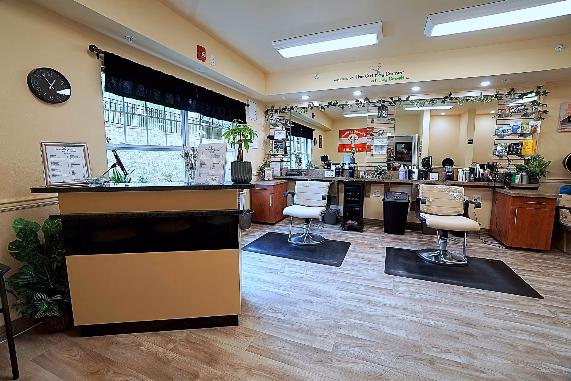 Interior view of a hair salon area in a retirement living facility with two salon chairs on black mats facing a large mirror. The room has light wood flooring, a reception desk with plants and menus, a clock on the wall, and decorative greenery above the mirror. The space is well-lit with ceiling lights and natural light from a window.
