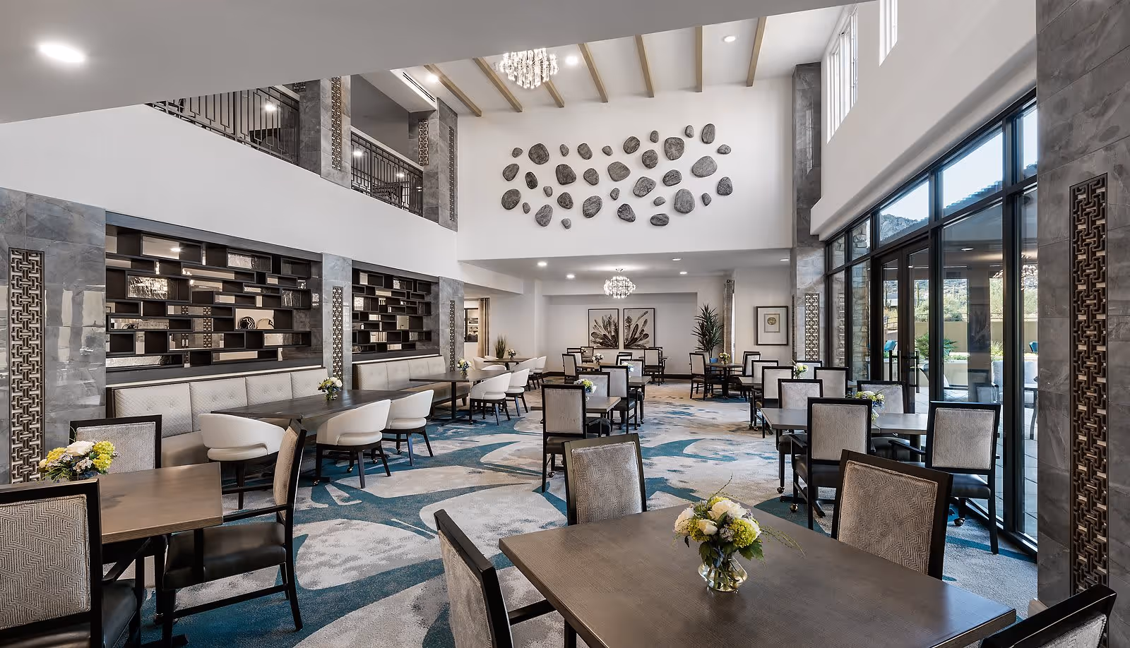 A spacious and modern dining room with multiple tables and chairs arranged neatly. The room features large windows on one side allowing natural light to brighten the space. There is a decorative wall with artistic stone pieces and a chandelier hanging from the ceiling. The carpet has a blue and white abstract pattern, and small flower arrangements are placed on the tables.