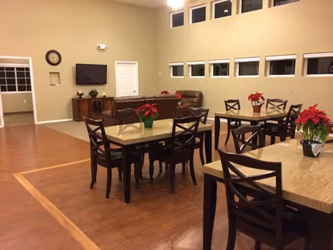 A spacious common area in a senior living facility with several wooden tables and chairs arranged on a wood floor. Each table has a red poinsettia plant as decoration. In the background, there is a seating area with brown leather sofas, a wall-mounted TV, a clock, and a wooden cabinet. The walls are beige with several small rectangular windows near the ceiling.