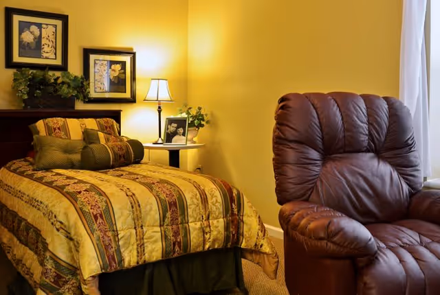 A cozy bedroom with a single bed covered in a yellow and brown patterned bedspread, a dark brown leather recliner chair, a small side table with a lamp, a framed photo, and a small plant. Two framed floral pictures hang on the yellow wall above the bed.