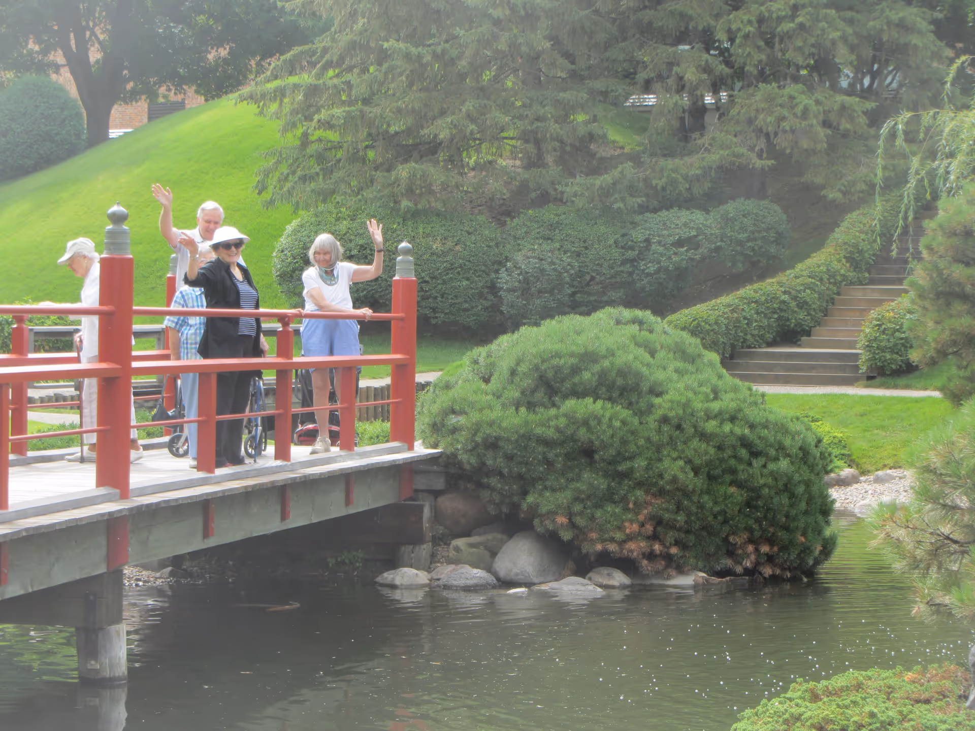 A group of elderly people standing on a red wooden bridge over a pond in a lush garden area. Some of them are waving and smiling. The background shows green bushes, trees, and a set of stairs leading up a grassy hill.