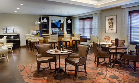 Communal lounge with round tables and leather chairs, a bar with upholstered stools, a piano and decorative artwork.