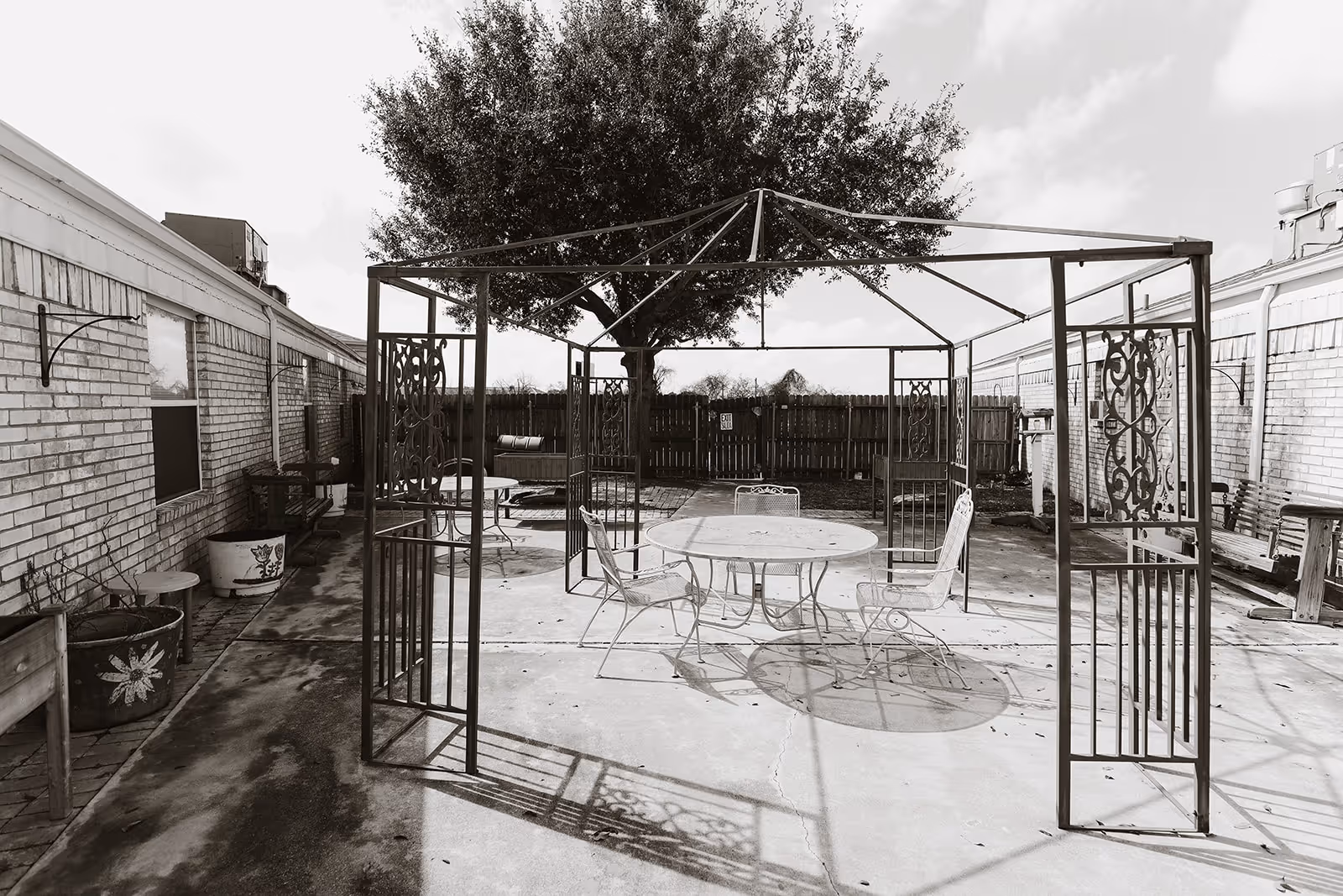 Outdoor patio area with a metal gazebo frame, a round table, and several chairs on a concrete surface. The patio is surrounded by brick walls on two sides and a wooden fence at the back. There is a large tree providing some shade and several planters and benches along the walls.
