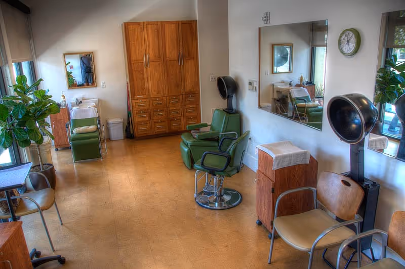 Interior of a senior living facility hair salon with green salon chairs, a hair dryer, wooden cabinets, a large mirror on the wall, and a potted plant near a window.
