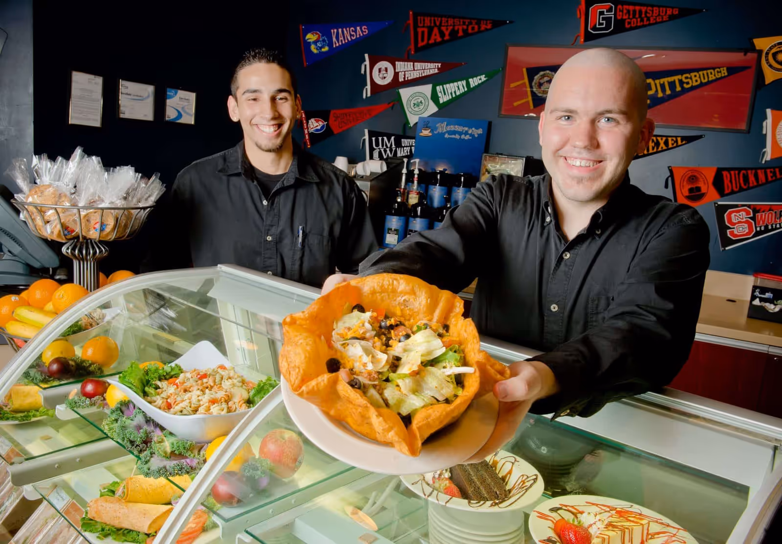 Two men behind a deli counter smiling, one man is holding out a taco salad in a crispy tortilla bowl. The counter displays various food items including pasta salad, fruits, and desserts. The background wall is decorated with multiple college pennants.