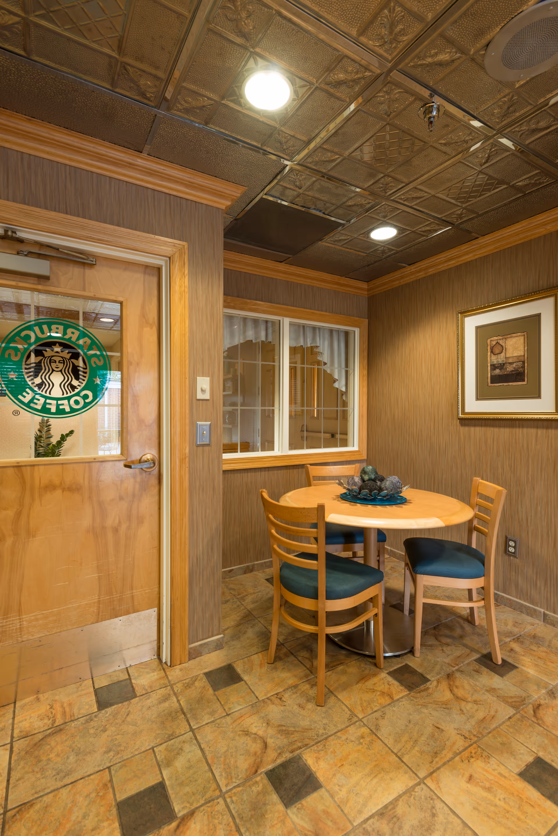 Small seating area with a round wooden table and three wooden chairs with green cushions in a room with brown paneled walls and tiled floor. A door with a Starbucks Coffee logo is visible on the left, and a framed picture hangs on the right wall. The ceiling has decorative metal tiles with recessed lighting.