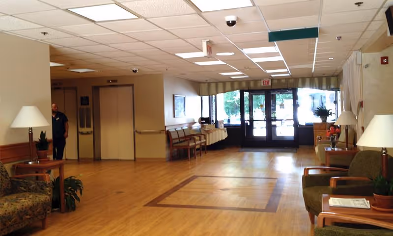Interior view of a senior living facility lobby area with wooden flooring, seating arrangements including chairs and small tables with lamps and plants, a person standing near two elevators on the left, and glass exit doors at the far end letting in natural light.