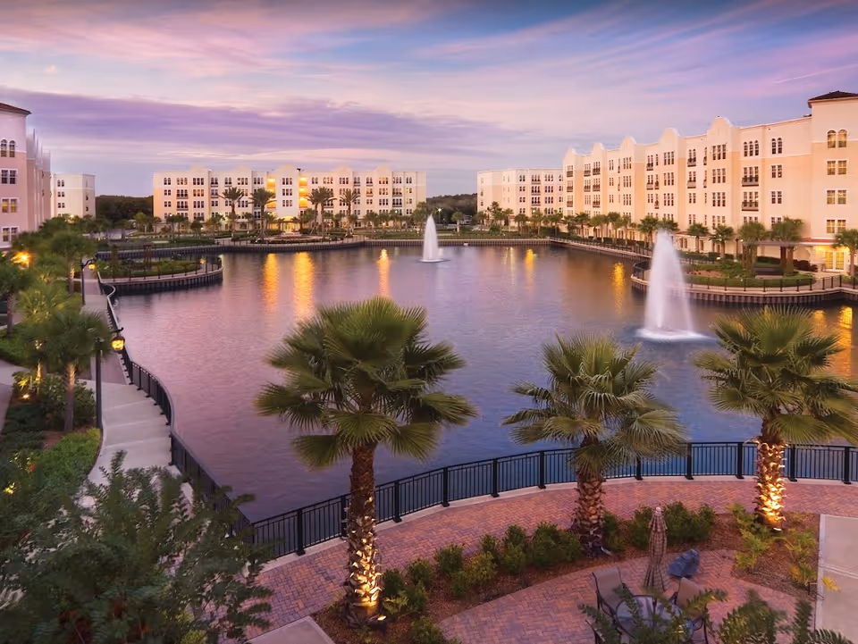 View of a senior living facility named The Glenridge on Palmer Ranch at dusk, featuring a large pond with two water fountains, surrounded by palm trees, landscaped walkways, and multi-story residential buildings with lights on.