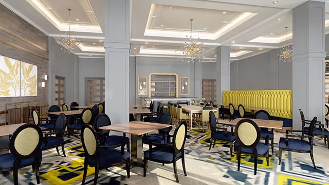 A spacious dining room with multiple wooden tables and black chairs with yellow cushions. The room features a modern design with gray walls, decorative ceiling lights, and a yellow cushioned bench along one wall. There is a patterned carpet with yellow, blue, and gray colors, and artwork on the wall.