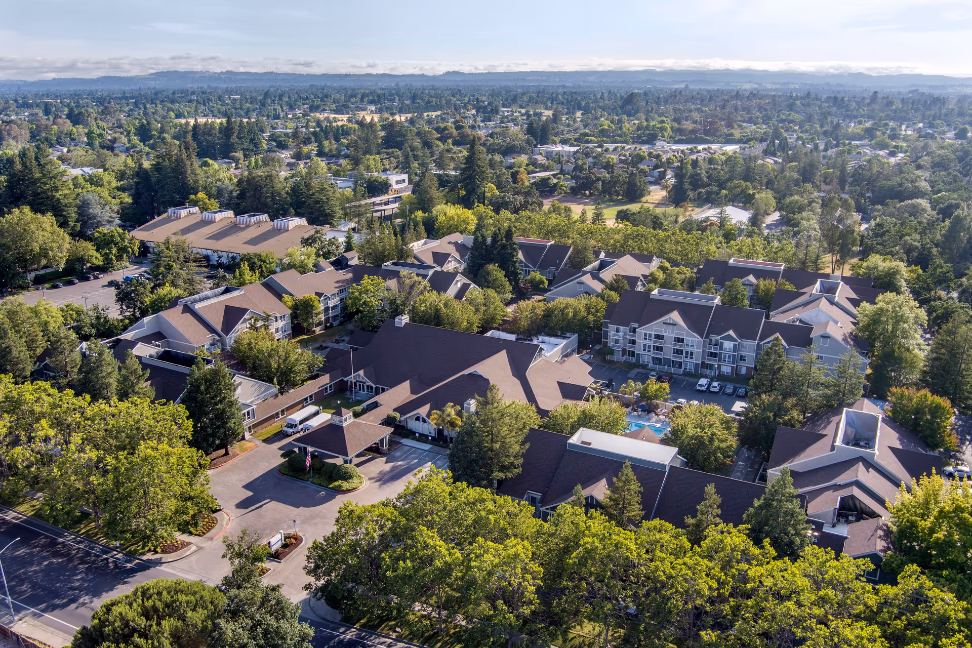Aerial view of Brookdale Paulin Creek senior living facility surrounded by trees and greenery, showing multiple buildings with brown roofs, parking areas, and a swimming pool, with a suburban landscape in the background under a partly cloudy sky.