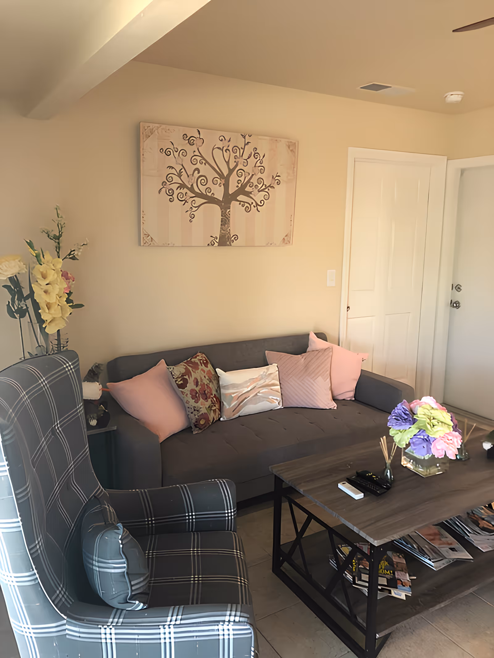 A cozy living room with a gray sofa adorned with pink and patterned pillows, a plaid armchair, a wooden coffee table with a glass vase of colorful flowers, and a wall art piece depicting a stylized tree. There is a door and a closet in the background.