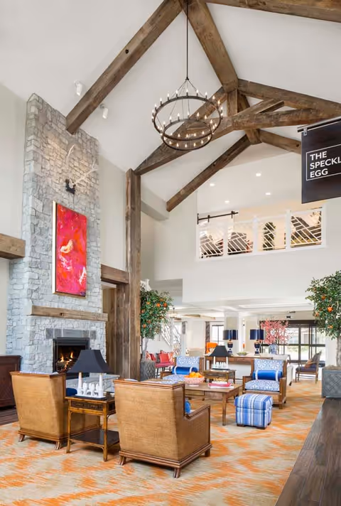 A spacious and bright living room area with high vaulted ceilings featuring exposed wooden beams and a large circular chandelier. The room has a stone fireplace with a colorful abstract painting above it. There are several comfortable chairs and sofas arranged around wooden coffee tables on an orange patterned carpet. Large windows and indoor plants add to the welcoming atmosphere.