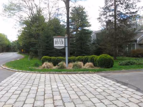 Stone-paved driveway leads to a front lawn with a sign for Glen River and trees.