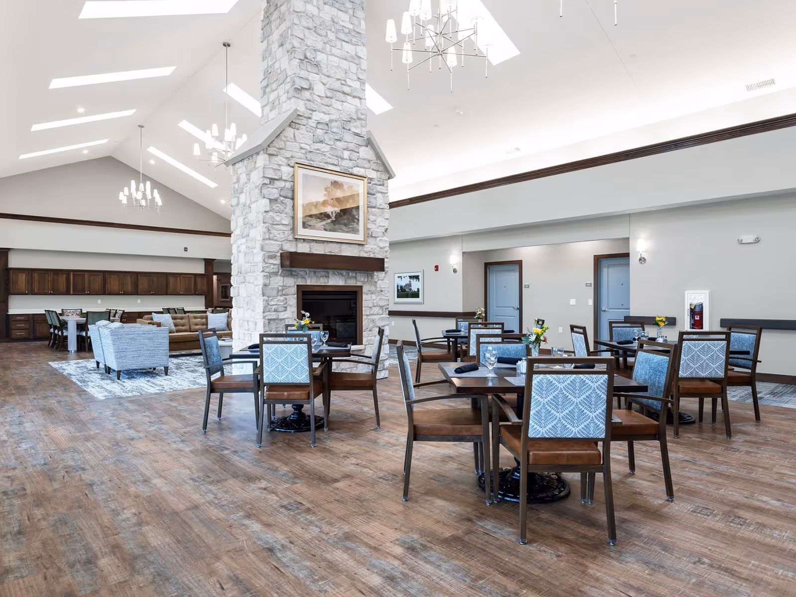 Spacious senior living dining area with wooden floors, multiple tables and chairs with blue patterned upholstery, a large stone fireplace in the center, and a seating area with sofas and armchairs in the background under a high ceiling with skylights and chandeliers.