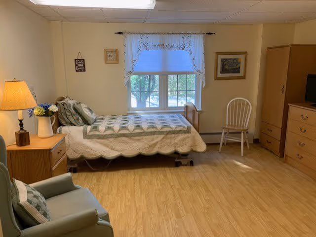 A cozy bedroom in a senior living facility featuring a single bed with a quilted bedspread, a wooden nightstand with a lamp and a vase of flowers, a comfortable armchair with a cushion, a wooden chair near a window with white curtains, and a wooden dresser with a TV on top. The room has light-colored walls and wood flooring.