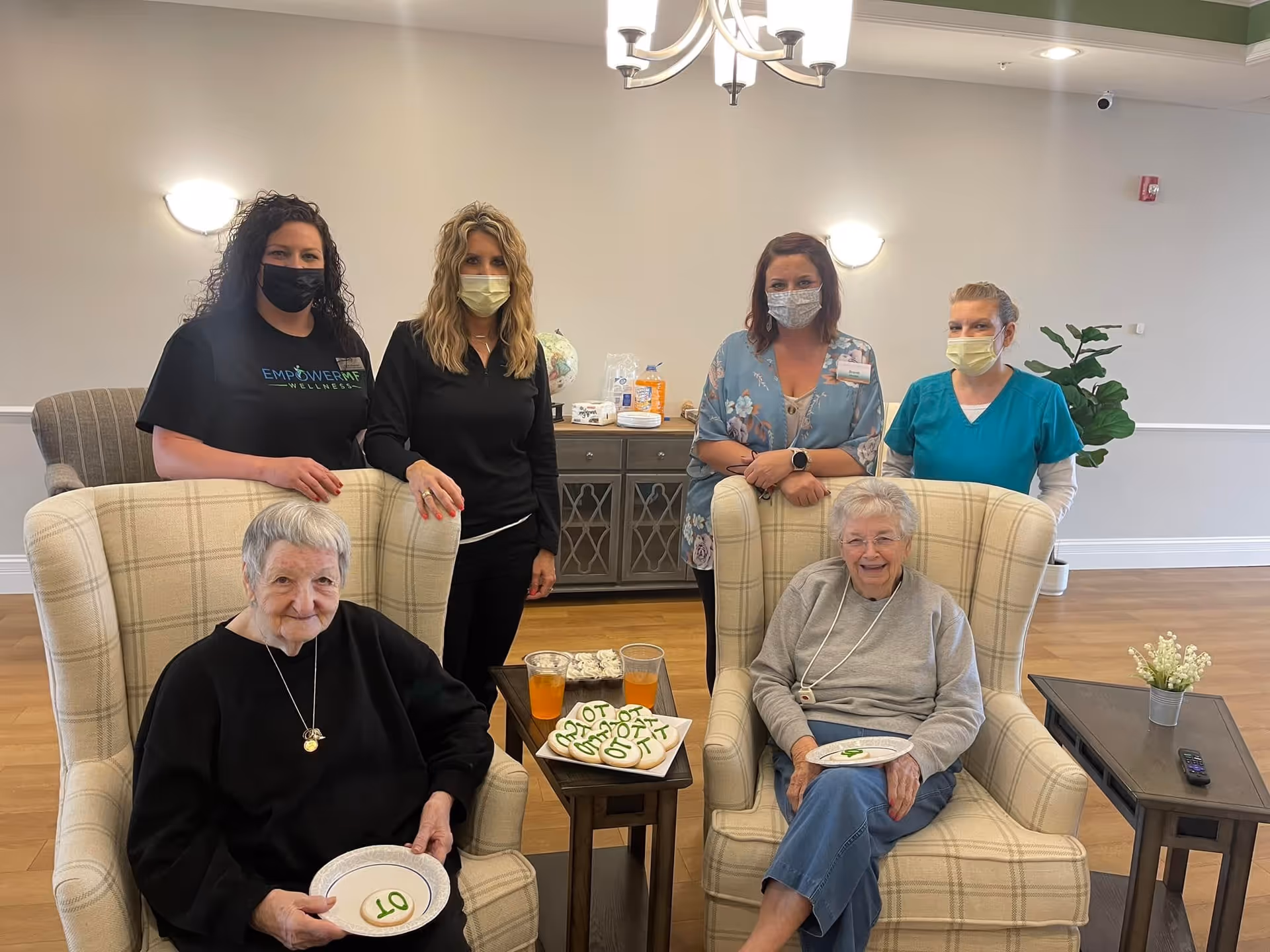 Two elderly women sit in plaid armchairs with plates and drinks on a small table while four masked staff stand behind them in a communal living room.