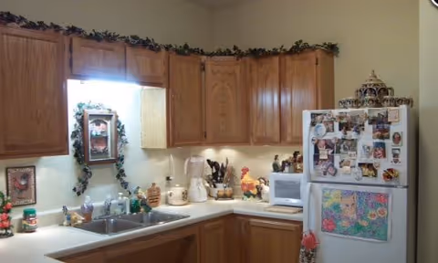 A kitchen with wooden cabinets, a white countertop, and a white refrigerator covered with various magnets and photos. The countertop has a blender, a microwave, and several decorative items. There is a small window above the sink with a mirror and greenery decoration around the top of the cabinets.