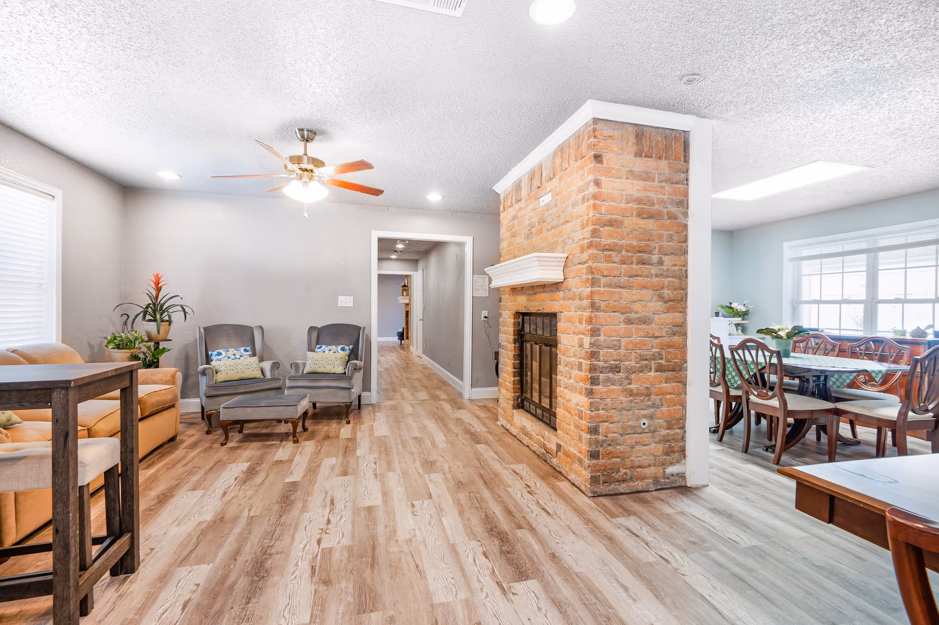 Bright communal living area with seating, a brick fireplace, and a dining table visible through an open doorway.