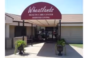 Entrance to Wheatlands Health Care Center Assisted Living with a maroon awning displaying the facility name. The entrance is covered and flanked by two large planters with greenery. The building exterior is light-colored brick with a clear pathway leading to glass doors.