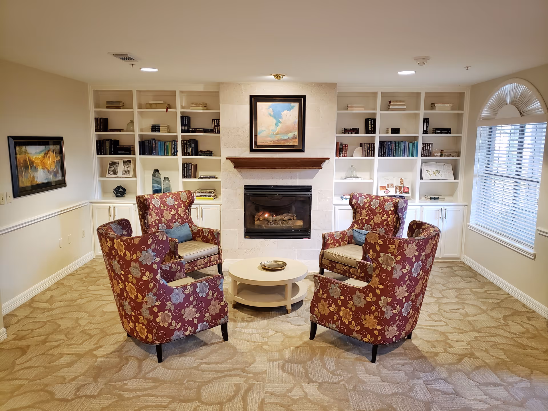 A cozy sitting area with four floral-patterned armchairs arranged around a round white coffee table. Behind the chairs is a fireplace with a painting above it, flanked by built-in white bookshelves filled with books and decorative items. A window with blinds is on the right side, and a framed artwork hangs on the left wall. The room has beige patterned carpet and soft lighting.
