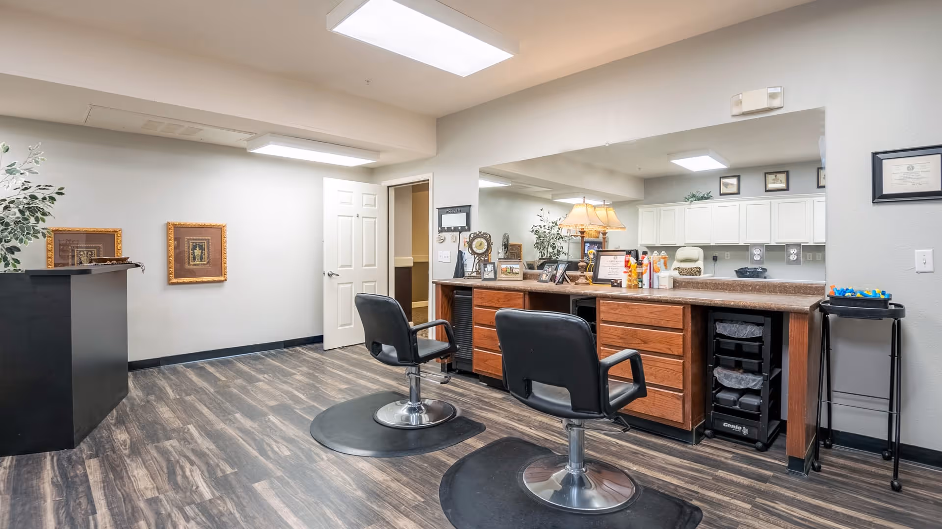 Interior view of a salon area in a senior living facility with two black salon chairs in front of a long wooden counter with drawers. The counter has various hair care products and a large mirror above it. The room has wood-patterned flooring, white walls, framed pictures, and a door leading to another room.