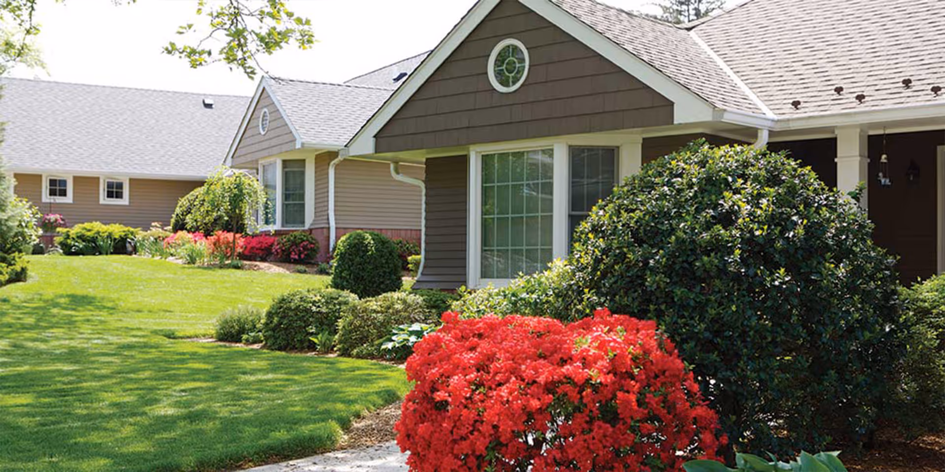 Front exterior of a single-story senior living building with a manicured lawn, shrubs, and bright red flowering bushes.