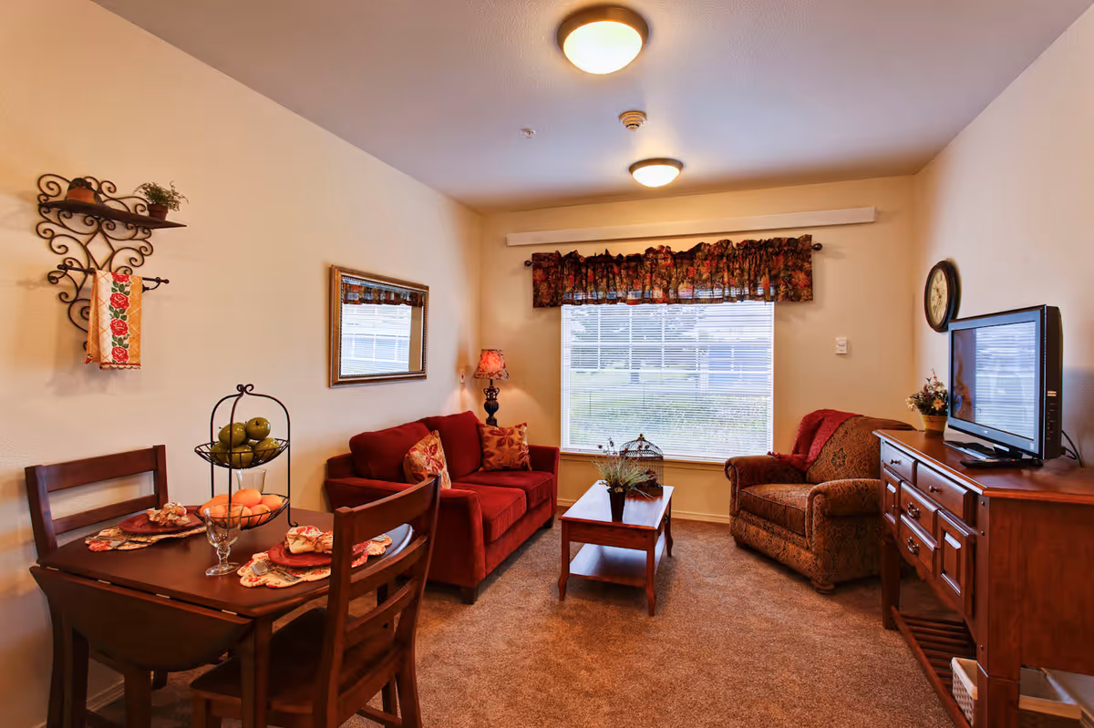 Cozy living room with a small dining table, red sofa, armchair, coffee table and TV in front of a large window.