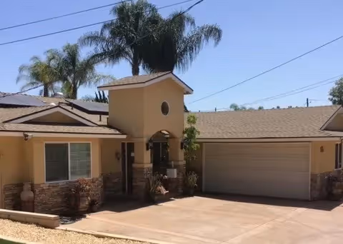 Single-story tan stucco building with a two-car garage, central arched entry tower, palm trees behind it, and a paved driveway.