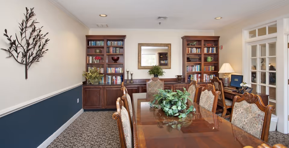 A well-lit interior room with a long wooden table surrounded by ornate chairs with patterned upholstery. On the table is a green leafy plant centerpiece. The back wall features two wooden bookshelves filled with books and decorative items, with a large framed mirror centered between them. To the right, there is a desk with a computer, lamp, and telephone. The walls are painted white with a dark blue lower half and decorated with a metal wall art piece shaped like branches with leaves.