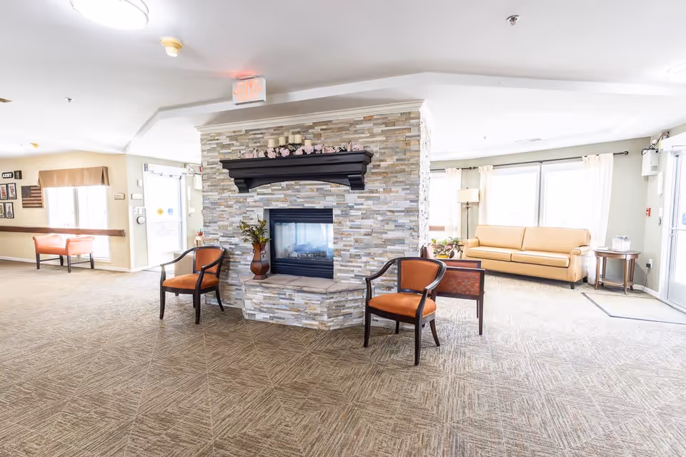 A bright and spacious common area in a senior living facility featuring a central stone fireplace with a black mantel decorated with candles and flowers. Two orange cushioned chairs are placed in front of the fireplace. The room has beige carpet flooring, large windows with light curtains, a beige sofa, a floor lamp, and a small side table near the entrance door.