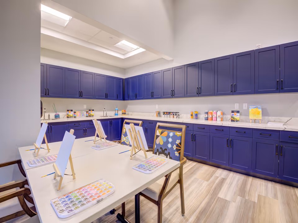 A bright art room with a large table set up for painting, featuring small easels with blank canvases and watercolor paint palettes. The room has purple cabinets lining the walls, a marble countertop with various art supplies, and patterned chairs around the table. The floor has light wood-style flooring and the ceiling has recessed lighting.