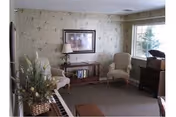 Cozy sitting room with two upholstered armchairs, a side table with lamp and framed artwork, a window, and decorative plants.